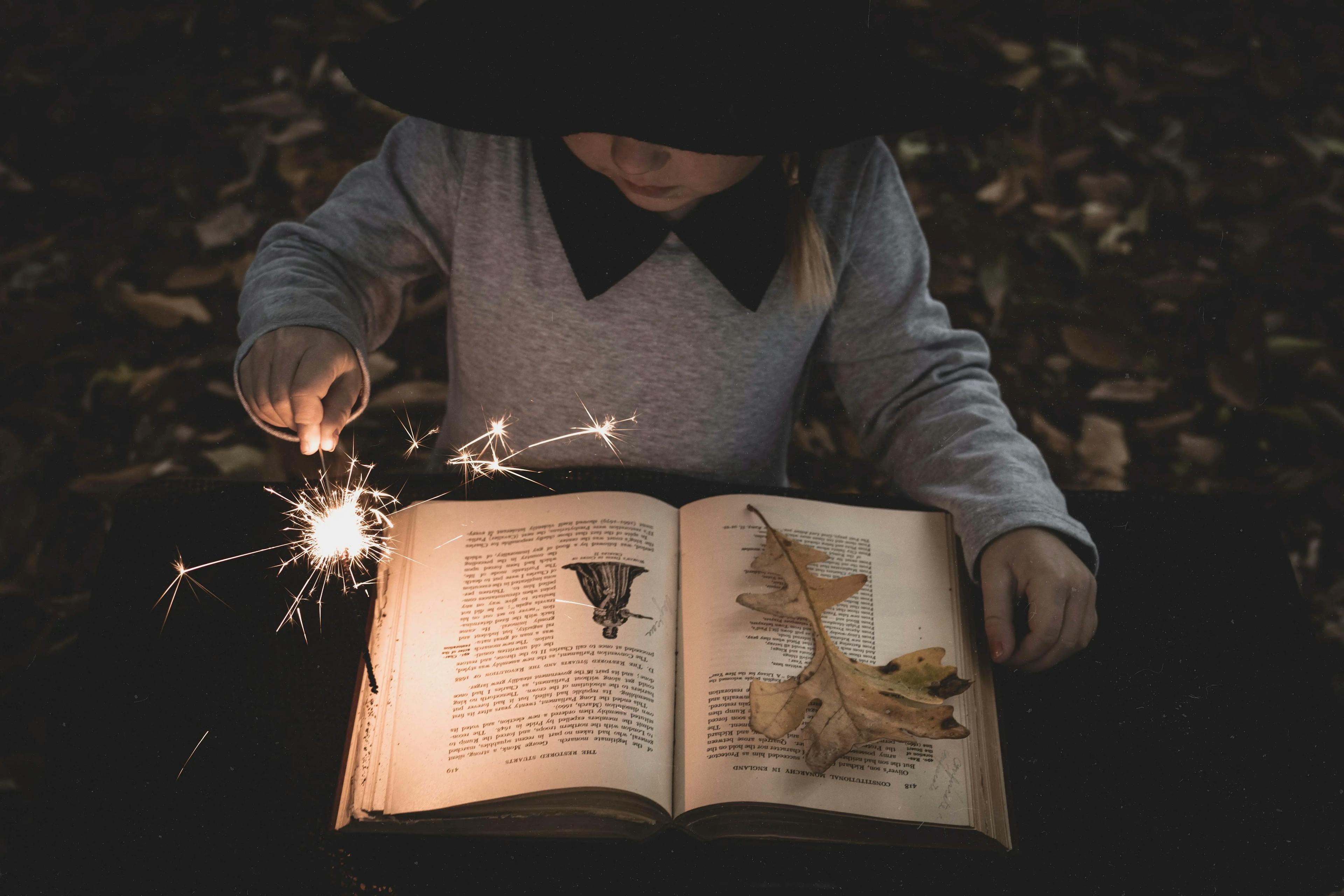A girl in a witch costume sits in the foliage with a book and a sparkler in her hands